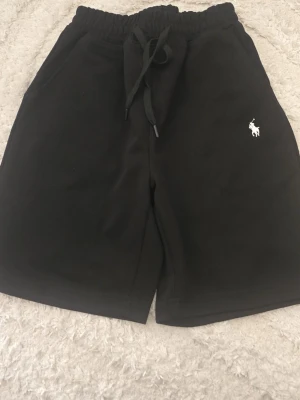 Svarta shorts Ralph Lauren - Svarta shorts från Polo Ralph Lauren med vit broderad logga på vänster ben. Mjuk bomullskvalitet, elastisk midja med snörning och två sidofickor. Perfekta för sommaren och chill dagar.