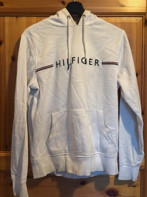 Tommy Hilfiger Hoodie - Vit hoodie från Tommy Hilfiger med broderad logga och tunna ränder i rött och marinblått över bröstet. Klassisk känguruficka framtill och mjukt bomullsmaterial. Perfekt för en avslappnad och stilren look. Behöver bara strykas. Använd ett par gånger. Pris kan alltid diskuteras💫💫