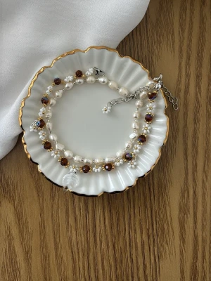 Pärlarmband med bruna kristaller - Unikt armband med vita pärlor, små blommor och bruna kristallpärlor. Armbandet har ett silverfärgat justerbart spänne och en genomskinlig hjärtberlock. Perfekt för dig som gillar detaljer och mixade material. 70kr/st