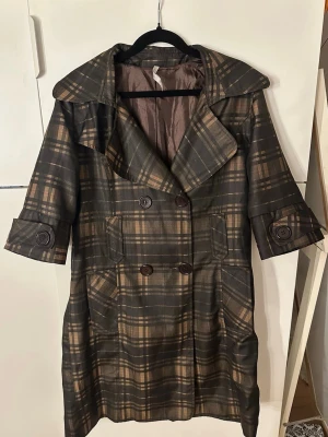 Rutig trenchcoat med 3/4 ärmar - Snygg trenchcoat i brunt och beige rutigt mönster med stora slag och dubbelknäppning. Jackan har trekvartsärmar med dekorativa knappar och två fickor framtill. Fodrad insida och markerad midja baktill. Storlek L men skulle säga mer åt S-M hållet