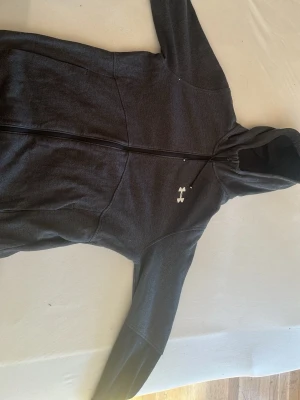 Mörkgrå hoodie från under armour - Mörkgrå hoodie från under armore med dragkedja hela vägen och vit logga på bröstet. Tröjan har huva och långa ärmar, samt en sportig och enkel design. Perfekt för dig som gillar outdoor eller vill ha en stilren hoodie till vardags.