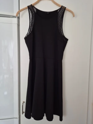 Klänning - Svart ärmlös klänning från H&M med transparenta meshdetaljer vid axlarna. Klänningen har en rund halsringning och en lätt utställd kjol som ger en snygg siluett. Perfekt för dig som gillar stilrena och enkla plagg.