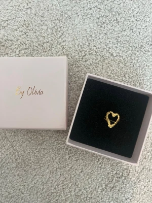Guldfärgad hjärtring By Olivia - Trendig guldfärgad ring från By Olivia med ett öppet hjärta som motiv. Ringen har en lite ojämn form. Ringen är justerbar och tål vatten!