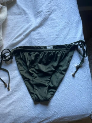 Mörkgröna bikiniunderdelar med knyt - Snygga mörkgröna bikiniunderdelar med knyt i sidorna. Klassisk modell med enkel design och tunna band som ger en trendig look. Perfekt för stranden eller poolen. Materialet känns mjukt och följsamt mot huden.