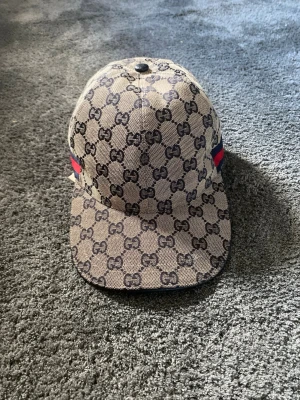 keps med GG-mönster - Snygg keps i beige med klassiskt GG-monogram över hela kepsen. På sidan finns en röd och blå rand som ger extra stilpoäng. Kepsen har böjd skärm och är tillverkad i canvasmaterial. Perfekt för dig som vill sticka ut med en lyxig accessoar.