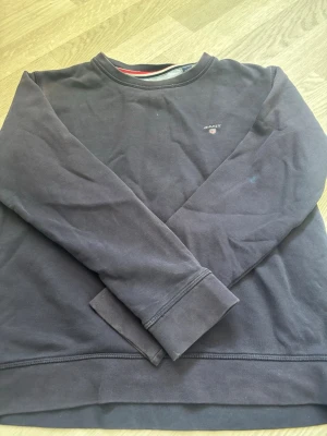 Mörkblå sweatshirt från Gant - Klassisk mörkblå sweatshirt från Gant med rund halsringning och diskret logga på bröstet. Tröjan har långa ärmar och ribbade muddar vid ärmslut och nederkant. Perfekt för en avslappnad och stilren look.