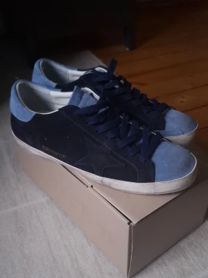 Golden Goose - Snygga Golden Goose Super-Star sneakers i mocka med mörkblå och ljusblå paneler. Klassisk stjärndetalj på sidan och vita sulor. Skorna har blå skosnören och är tillverkade i Italien. Perfekta för dig som gillar en stilren men ändå unik look.