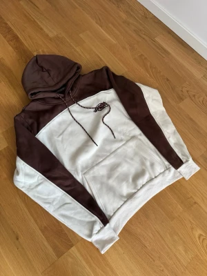 Brun och vit hoodie från Syna world - Snygg hoodie i brunt och vitt från syna world med broderad logga på bröstet. Hoodien har huva och coola kontrastdetaljer. Perfekt för dig som gillar streetwear och vill sticka ut lite extra.