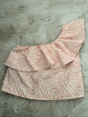 Zebramönstrad oneshoulder topp MNG - Snygg oneshoulder topp från MNG med rosa och vitt zebramönster. Toppen har volangdetalj över axeln och är tillverkad i ett lätt, glansigt material. Perfekt för dig som vill sticka ut med en trendig och lekfull stil.
