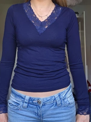 Mörkblå långärmad topp med spets - Mörkblå långärmad topp med djup V-ringning och dekorativ spetskant vid halsen och ärmsluten. Figurnära passform och stretchigt material som sitter snyggt på kroppen. Perfekt att styla med jeans eller kjol.