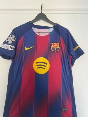 FC Barcelona matchtröja Nike - Snygg FC Barcelona matchtröja från Nike i blått och rött med gula detaljer. Tröjan har klubbmärke, Nike-logga och Spotify som sponsor på bröstet. Champions League-märke på ärmen. Tillverkad i lätt och ventilerande material, perfekt för fotbollsträning eller match.