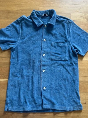 Blå frottéskjorta med kort ärm - Säljer en blå skjorta från Weekday i mjukt frottématerial. Skjortan har korta ärmar, klassisk krage, vita knappar och en bröstficka. Perfekt för dig som gillar en avslappnad och cool stil.