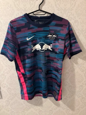 RB Leipzig träningströja Nike blå - Snygg RB Leipzig träningströja från Nike i blått med mönster i rosa, lila och turkos. Tröjan har klubbmärke och Nike-logga på bröstet samt Red Bull-tryck. Tillverkad i lätt och ventilerande syntetmaterial, perfekt för träning eller match.