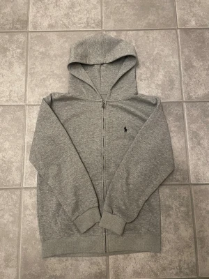 Grå zip-hoodie från Polo Ralph Lauren - Snygg gråmelerad hoodie med dragkedja från Polo Ralph Lauren. Klassisk design med huva, ribbade muddar och liten broderad logga på bröstet. Perfekt för en chill och stilren look. Mjuk bomullsblandning som känns skön mot huden.