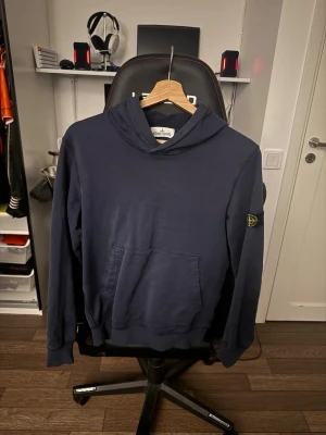 Mörkblå hoodie från Stone Island - Snygg mörkblå hoodie från Stone Island med klassisk patch på vänster ärm. Hoodien har en stor känguruficka framtill. Tillverkad i mjuk bomull för skön känsla och avslappnad stil. 