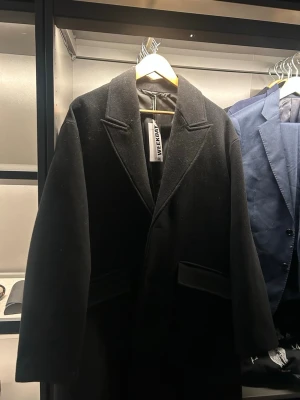 Svart ullkappa från Weekday - Stilren svart kappa från Weekday med klassisk krage och raka linjer. Kappan har två fickor framtill och är tillverkad i ett ullmaterial som ger en lyxig känsla. Perfekt för dig som vill ha en tidlös och trendig look.