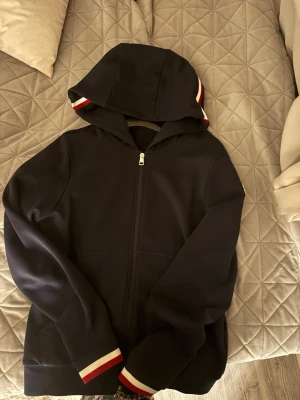 Marinblå Moncler hoodie med dragkedja - Snygg marinblå hoodie från Moncler med dragkedja framtill och huva. Detaljer i rött, vitt och blått på muddar och huva samt Moncler-logga på ärmen. Perfekt för dig som gillar exklusiva streetwear-plagg med stilrena detaljer.