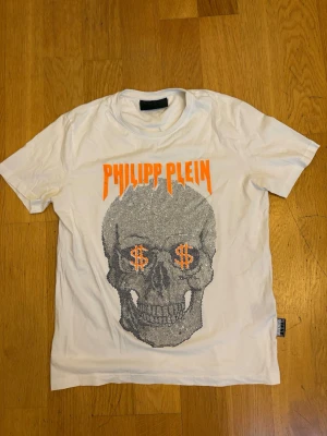Vit Philipp Plein t-shirt med döskalle - Cool vit t-shirt från Philipp Plein med ett stort döskallemotiv i silver och orange på framsidan. Loggan är tryckt i orange ovanför motivet och t-shirten har korta ärmar. Materialet är mjuk bomull och passformen är normal.