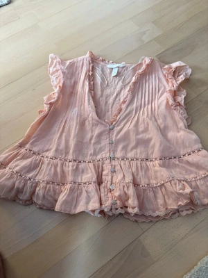 Persikofärgad blus med volanger H&M - Supersöt persikofärgad blus från H&M med volanger och spetsdetaljer. Blusen har knappar framtill, v-ringning och korta volangärmar. Tillverkad i ett tunt, luftigt material som känns lätt och somrig. Perfekt för dig som gillar romantisk och bohemisk stil.
