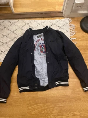 Svart bomberjacka med vita detaljer - Snygg svart bomberjacka från Gant med vita ränder på muddar och krage. Jackan har dragkedja, tryckknappar och en bröstficka med dragkedja. Klassisk college-look och perfekt för dig som gillar sportig stil.