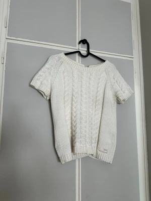 Vit kabelstickad topp Odd Molly - Säljer en cream vit kabelstickad topp från Odd Molly med korta ärmar och bred ribbad kant vid hals och nederkant. Tröjan har ett klassiskt flätmönster framtill och en liten Odd Molly-label nedtill. Prefekt när som och jättesnygg med en långärmad tröja under!