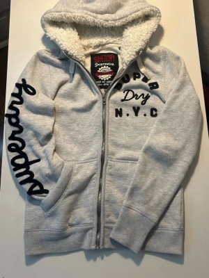 Grå Superdry hoodie med teddyfoder - Snygg ljusgrå hoodie från Superdry med dragkedja, svart broderad logga på ärmen och tryck på bröstet. Insidan av huvan är fodrad med mjuk teddy för extra komfort. Perfekt för en chill och avslappnad stil. Står M men passar även S