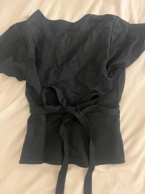 Svart omlottopp med knytband - !!Om du köper välj helst att hämta vid upphämtningsställe då det blir enklare för mig att posta på det viset!!                                      Säljer en svart omlottopp med korta ärmar och breda knytband i midjan som ger en snygg siluett. Toppen har en enkel, stilren design och är perfekt att styla med jeans eller kjo