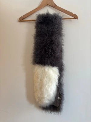 Fluffig grå och vit fuskpäls halsduk - Supermjuk och fluffig halsduk i fuskpäls med grå och vit färgblock. Insidan är fodrad med slätt tyg för extra komfort. Perfekt accessoar för att lyfta din vinterstil och hålla dig varm under kalla dagar.