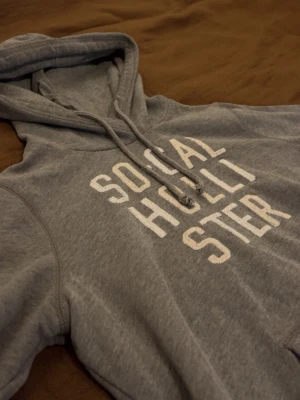 Hollister hoodie - Snygg grå hoodie från Hollister med vit text 'SOCAL HOLLISTER' på bröstet. Klassisk modell med huva och dragsnören samt en stor magficka. Tillverkad i mjukt material, perfekt för chill dagar.