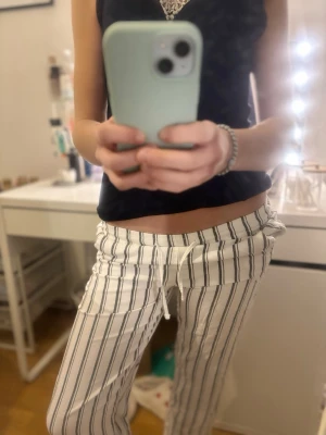 Vita randiga loungewear-byxor - Säljer ett par vita loungewear-byxor med svarta vertikala ränder och dragsko i midjan. Byxorna har en avslappnad passform och är perfekta för chill hemma. Materialet känns mjukt och bekvämt, perfekt för mysiga dagar.