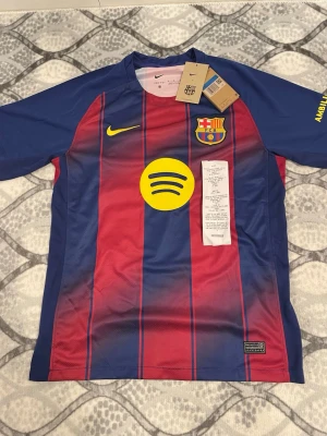 Lamine yamal fc Barcelona tröja - Säljer en officiell FC Barcelona matchtröja från Nike med klassiska röda och blå vertikala ränder. Tröjan har klubbmärke, Nike-logga och Spotify som sponsor i gult på bröstet. Tillverkad i lätt och ventilerande material, perfekt för fotboll eller träning.