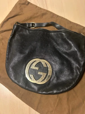 Svart läderväska från Gucci - Gucci axelremsväska för kvinna i svart läder. Väskan har en rundad form med justerbar axelrem och dragkedja som stängning. Framsidan pryds av en stor logodetalj. Invändigt finns ett huvudfack med mindre fack för organisering av personliga tillhörigheter. Passar för vardagsbruk och för att bära nödvändigheter som plånbok, mobil och nycklar. Levereras med tygpåse från Gucci enligt bild. Inköpt i Paris, kvitto finns ej kvar. Måttet är 36 x 29 cm.