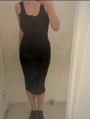 Svart ärmlös klänning med knappar - Snygg svart ärmlös klänning med knappar framtill. Klänningen har en kroppsnära passform och går ner till knäna. Perfekt för dig som gillar en enkel men stilren look.