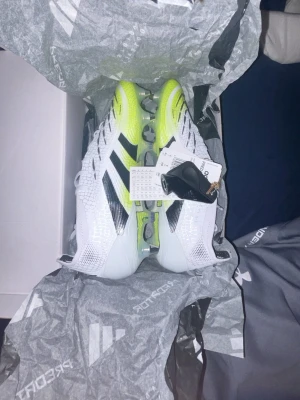 Adidas Predator fotbollsskor vit/gul - Säljer ett par ”Helt nya” Adidas Predator fotbollsskor i vitt och neongult med svarta detaljer. Skorna har en modern design med texturerad ovandel och metallfärgade dobbar för extra grepp. Tillverkade i syntetmaterial och levereras i originalkartong.