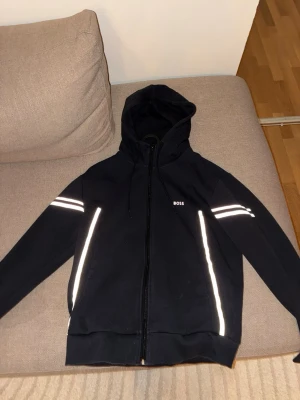 Svart hoodie med reflexdetaljer från BOSS - Snygg mörkblå hoodie från BOSS med dragkedja, huva och bruda sträck med när man tar bild så blir det vita  reflexdetaljer längs ärmar och fickor. Liten BOSS-logga på bröstet. Perfekt för dig som vill ha en stilren men ändå sportig look. Tillverkad i mjukt material för extra komfort. PRIS GÅR Å DISKUTERA VID SNABB AFFÄR 