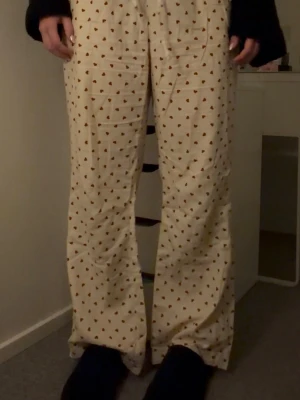 Vita pyjamasbyxor med hjärtmönster - Säljer ett par vita pyjamasbyxor med sött rött hjärtmönster. Byxorna har rak passform och band i midjan som går att justera. Perfekta för mysiga hemmakvällar och loungewear. Materialet känns mjukt och skönt mot huden.