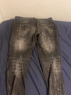 Svarta Dsquared2 jeans - Säljer ett par svart/gråa Dsquared2 jeans med coola slitningar, färgstänk och distressed detaljer. Jeansen har raka ben, fem fickor och en tydlig Dsquared2-logga vid gylfen. Materialet är klassisk denim med en trendig, sliten look.