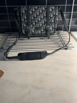 Christian Dior axelväska med monogram - Snygg axelväska från Christian Dior med klassiskt grått och svart monogrammönster. Väskan har justerbar axelrem med vadderad del, två spännen framtill och innerfack med dragkedja. Materialet är canvas och läder.