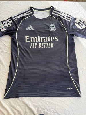 Real Madrid bortatröja Adidas - Mörkblå Real Madrid bortatröja från Adidas med vita detaljer och klubbmärke på bröstet. Tröjan har rund hals, tre vita ränder på axlarna och sponsorn Emirates Fly Better tryckt framtill. Tillverkad i lätt och ventilerande material, perfekt för fotboll.