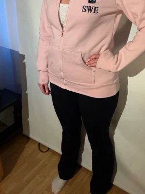 Ljusrosa hoodie med tryck SWE - Ljusrosa hoodie med dragkedja och fickor framtill. Tryck med texten SWE på bröstet och HORSE längs ena ärmen i mörkblått. Klassisk huva och ribbade muddar. Perfekt för dig som gillar en sportig och avslappnad stil.