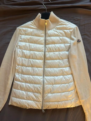 Beige quiltad cardigan från Moncler - Snygg beige cardiganjacka från Moncler med quiltad, glansig front och rygg i dunmaterial samt stickade ärmar och sidor. Dragkedja framtill och Moncler-logga på ärmen. Hög krage och stilren design som passar perfekt till lager-på-lager.