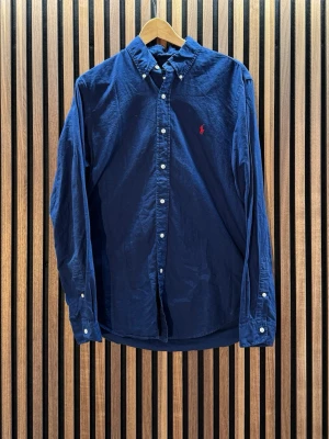 Marinblå skjorta från Ralph Lauren - Klassisk mörkblå skjorta från Ralph Lauren. M i storlek. Skick 9/10. (Har små vita sträck som ser ut att gå bort i tvätten, se bild nr 7) Hör av er vid funderingar. Pris ej hugget i sten