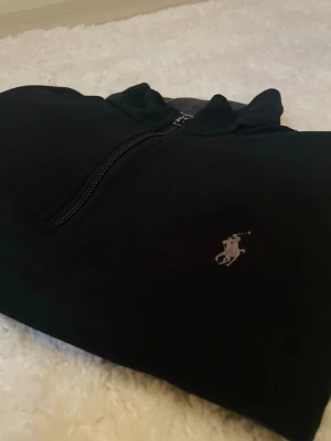 Svart halvzip från Ralph Lauren - Svart långärmad tröja från Ralph Lauren med halvzip och klassisk broderad logga på bröstet. Tröjan har ståkrage med grå insida och är tillverkad i mjukt material, perfekt för en stilren och avslappnad look.