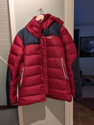 Röd dunjacka från Bergans of Norway - Snygg röd dunjacka från Bergans of Norway med svarta detaljer på axlar och ärmar. Jackan har huva, dragkedja framtill och två stora fickor med vita dragkedjor. Perfekt för kalla vinterdagar och riktigt varm tack vare dunfyllningen.
