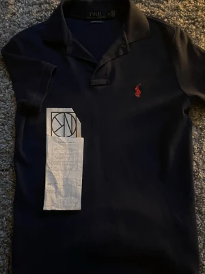 Mörkblå pikétröja från Ralph Lauren - Snygg mörkblå pikétröja från Ralph Lauren med klassisk krage och röd broderad logga på bröstet. Perfekt för en stilren look och enkel att matcha med jeans eller shorts. Custom slimfit i storlek Xs köpt på NK, sommarn 2024. Orginal pris 1499