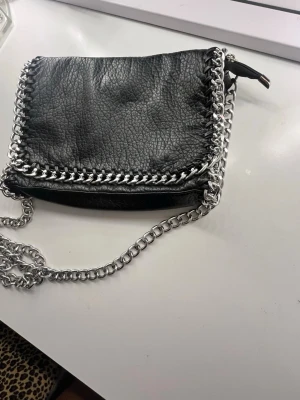  En Svart TIAMO ÄKTA axelväska med kedjedetaljer  - Snygg svart tiamo i silverfärgade kedjor längs kanterna och som axelrem. Väskan har en dragkedja på baksidan och cool struktur i materialet. Äkta skin Perfekt för dig som gillar en edgy stil. Köpte för 1500kr säljer för 800kr🥰
