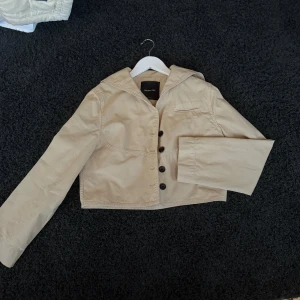 Beige kort jacka från Massimo Dutti - Snygg beige jacka från Massimo Dutti med croppad passform och klassisk krage. Jackan har långa ärmar, knäppning framtill med svarta knappar och stilrena sömdetaljer. Perfekt för dig som vill ha en trendig och enkel look.