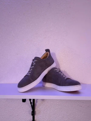 Christian Louboutin Grå - Stilrena gråa Christian Louboutin sneakers i mocka med vita sulor och röda undersidor. Skorna har klassisk låg profil, vita skosnören och diskreta detaljer. Perfekta för dig som gillar minimalistisk design och vill sticka ut med en färgglad sula. - Dom är endast använd en helg på kreta så dom är som ny! 