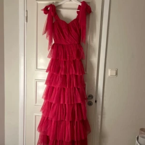 Rosa volangklänning med knytband - Snygg långklänning i starkt rosa med flera lager volanger och breda axelband som knyts i rosetter. Klänningen har omlottliv och är tillverkad i ett tunt, skirt material som ger en luftig känsla. Perfekt för dig som vill ha en elegant balklänning 