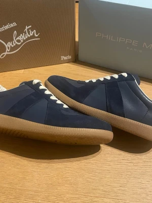 Mörkblå sneakers från Maison Margeila - Snygga mörkblå sneakers från Maison Margiela med ovandel i mocka och läder, vita skosnören och klassisk gummisula i brunt. Perfekt för dig som gillar stilrena och tidlösa sneakers med en sportig touch.